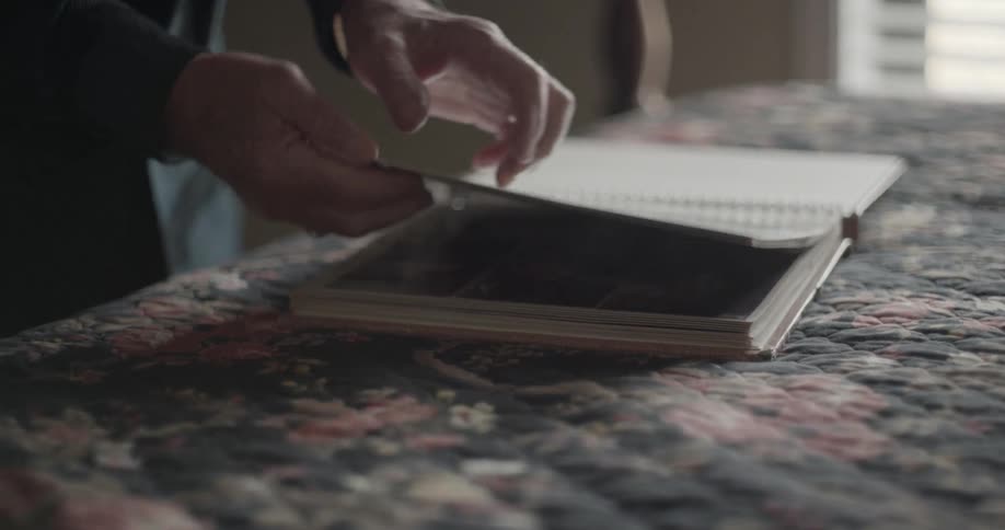 An older man is placing a picture album on a bed and leafing through the pages