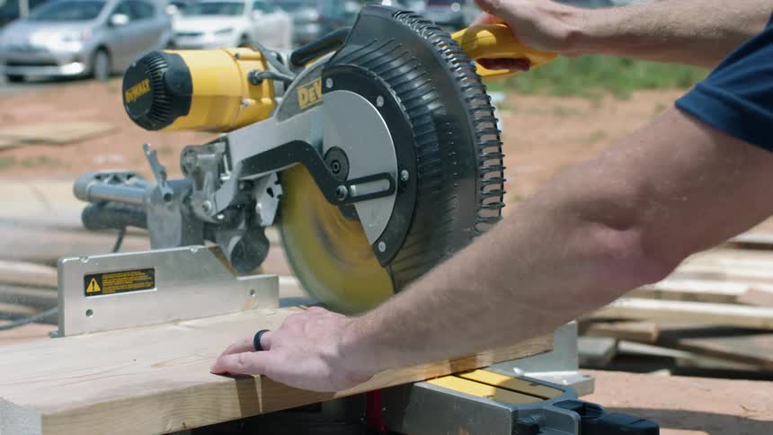 A man is using a table saw to cut a piece of wood outside during the day