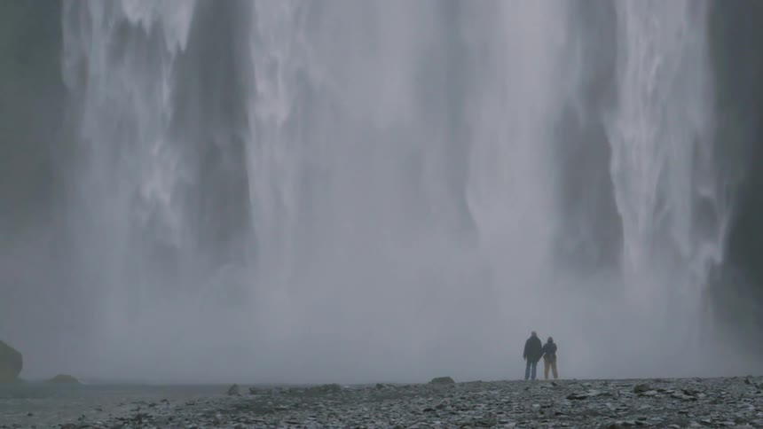 Two people standing at the foot of a huge waterfall hold hands