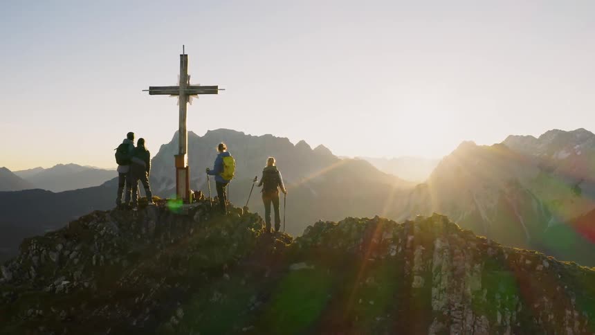 Hikers stand next to a cross on a mountain peak at sunset
