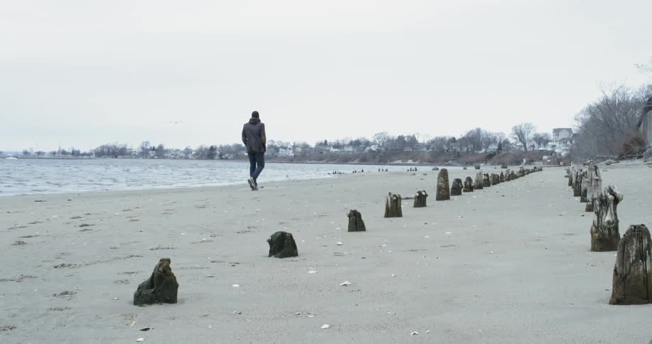A man wearing a jacket is walking along the beach on a cloudy day