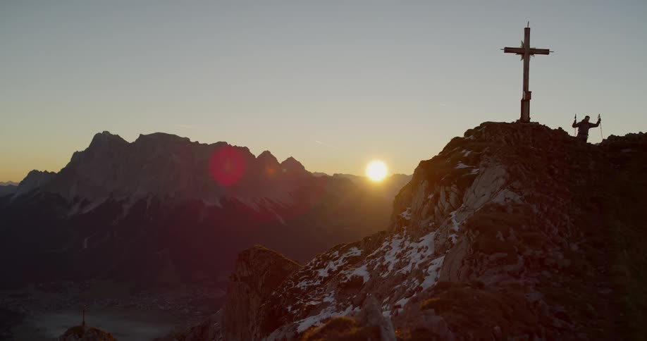 Sun setting behind mountains while a cross stands high in the foreground and people walking up to it