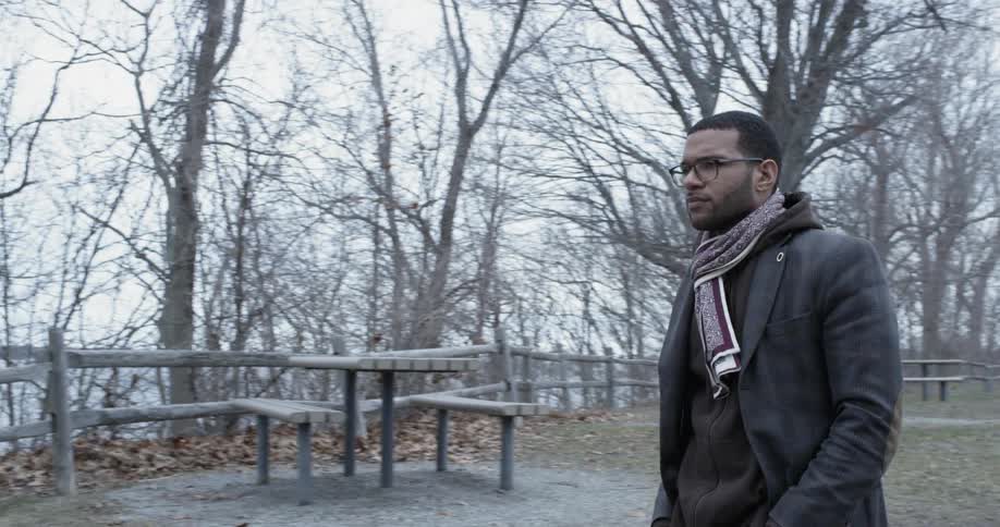 A man walks past trees and picnic tables in a park
