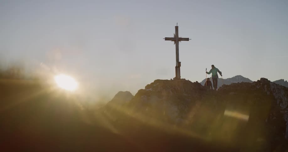 Hikers climb up to a cross on a mountain peak at sunset