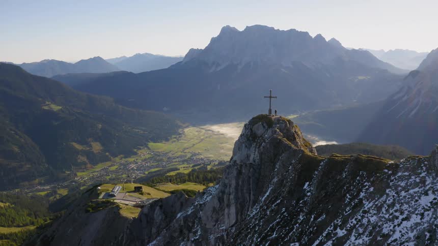 A large mountain area with snow and grass on the mountains and people standing nearing a cross on top of one of the mountains