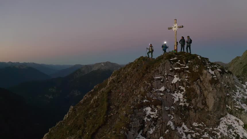 Four people stand near a cross on a mountain peak at dawn