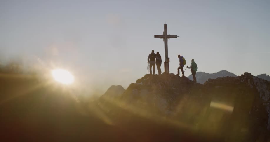 Four people stand near a cross on a mountain peak at dawn