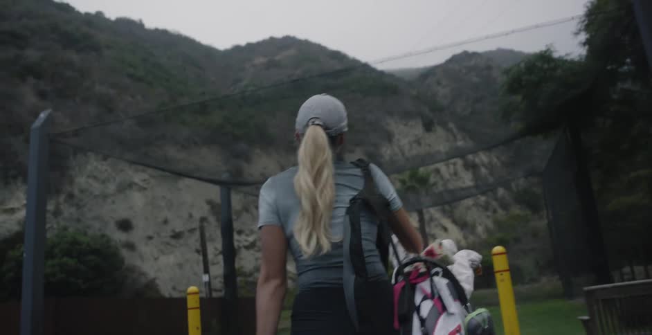 A blonde woman is carrying a golf bag and a bucket of golf balls