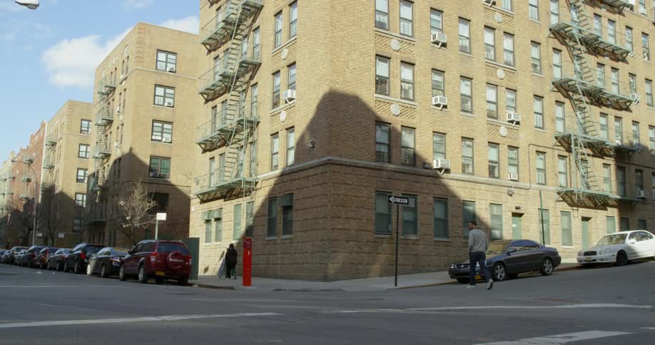 Two people walk on the sidewalk near a large apartment building in a city