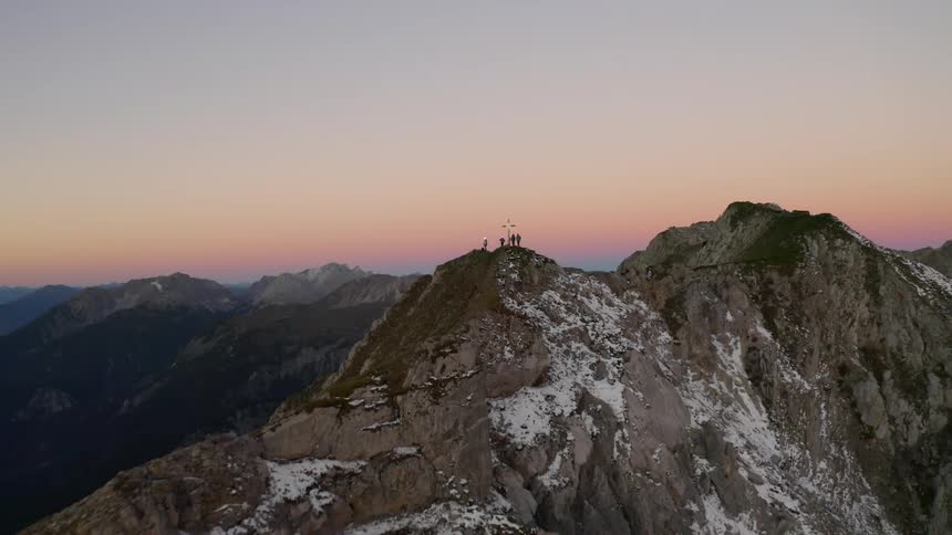 Hikers stand next to a cross on a mountain peak at sunset
