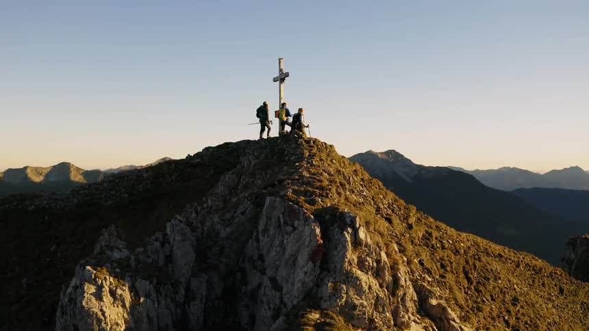 Hikers stand next to a cross on a mountain peak at sunset