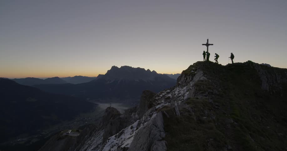 Hikers stand next to a cross on a mountain peak at sunset