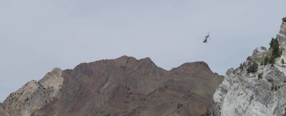 A helicopter carrying a cable and bucket flies toward a rocky ridge