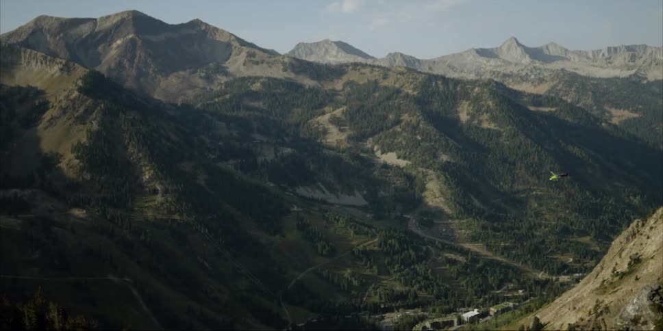 A helicopter is towing a bucket on a tether above a mountain valley in daylight