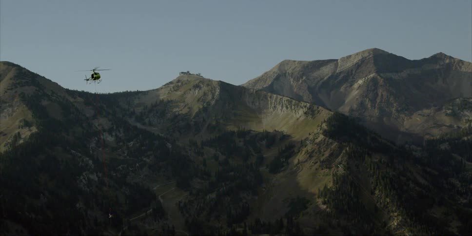 A helicopter dangling a tether is rising into the air above a mountain in daylight