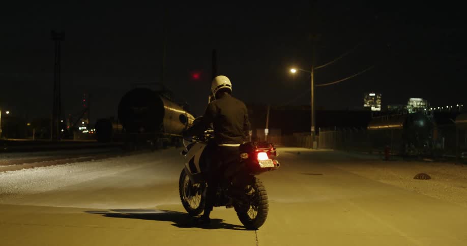 A man sits on a motorcycle at night in a city with a red laser dot floating around near his head