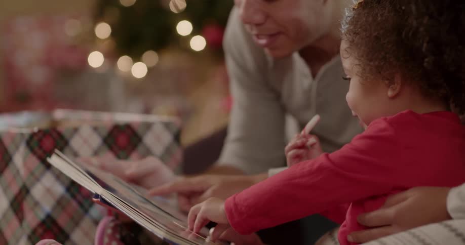 A man reads a young girl a book next to a christmas tree