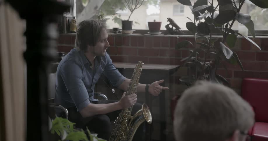 A man holding a saxophone is talking and gesturing as he sits in a room