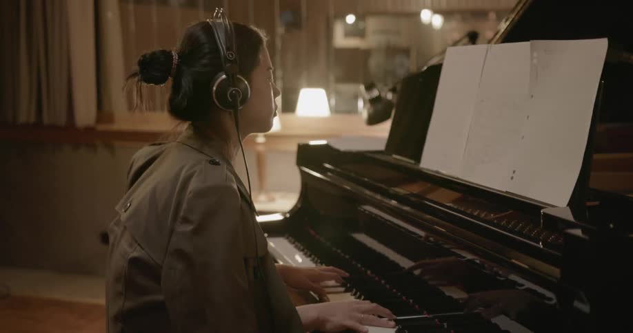 A woman sits at a piano in a room and plays while wearing headphones