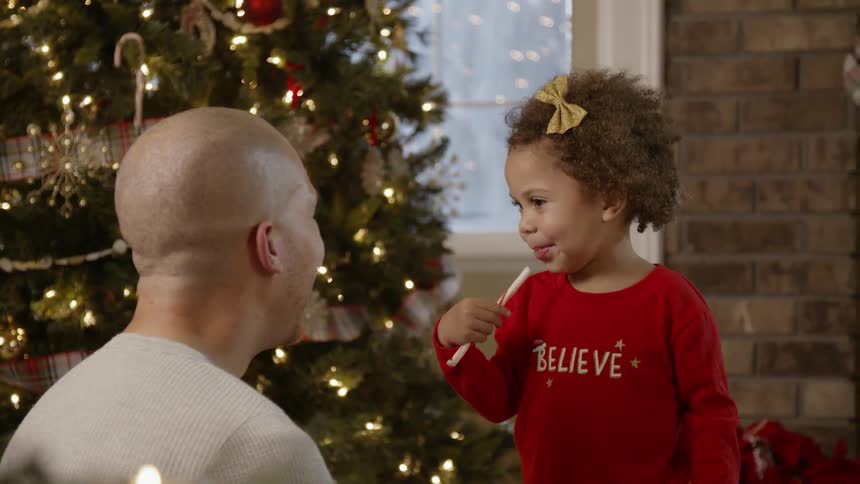 A girl licks a candy cane then offers it to her dad on christmas