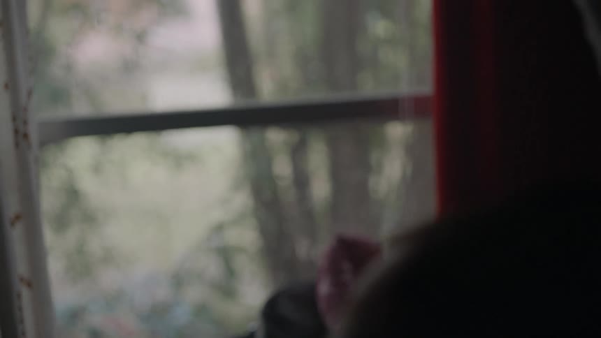 Rain is gently falling against a window and a woman is looking out of it