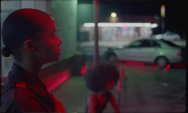 Two women stand on a city curb at night and one blows bubbles with chewing gum