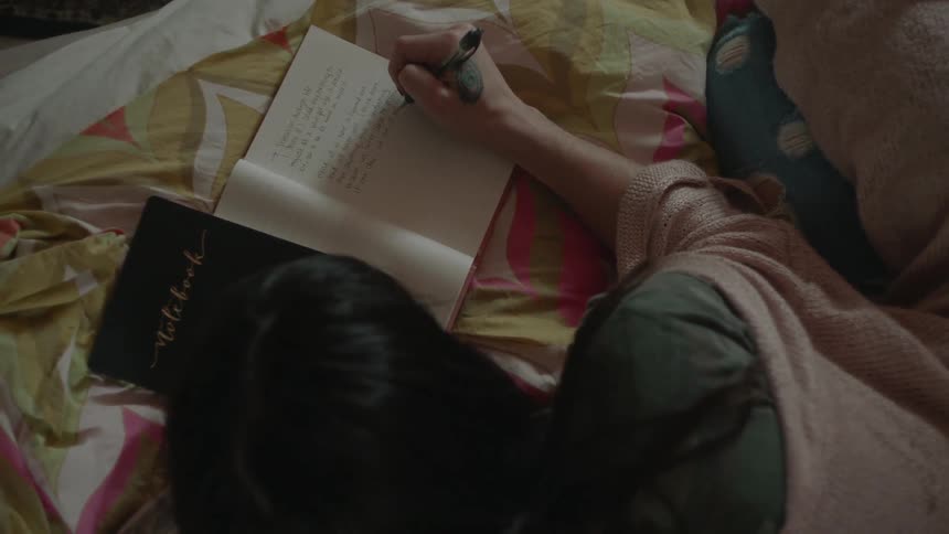 A woman is writing in a journal while sitting on a bed