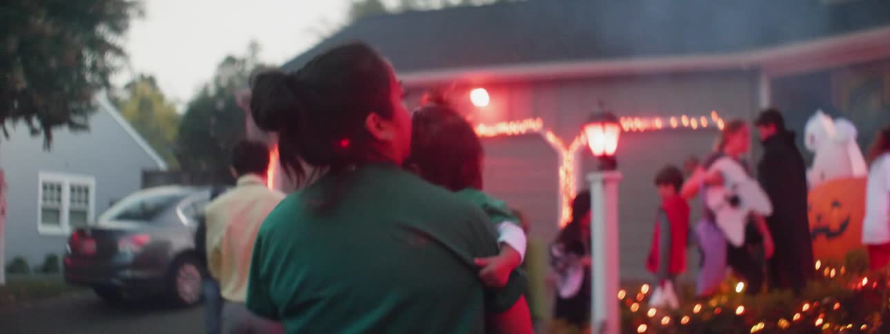 A woman is carrying a toddler as they trick-or-treat on halloween at dusk