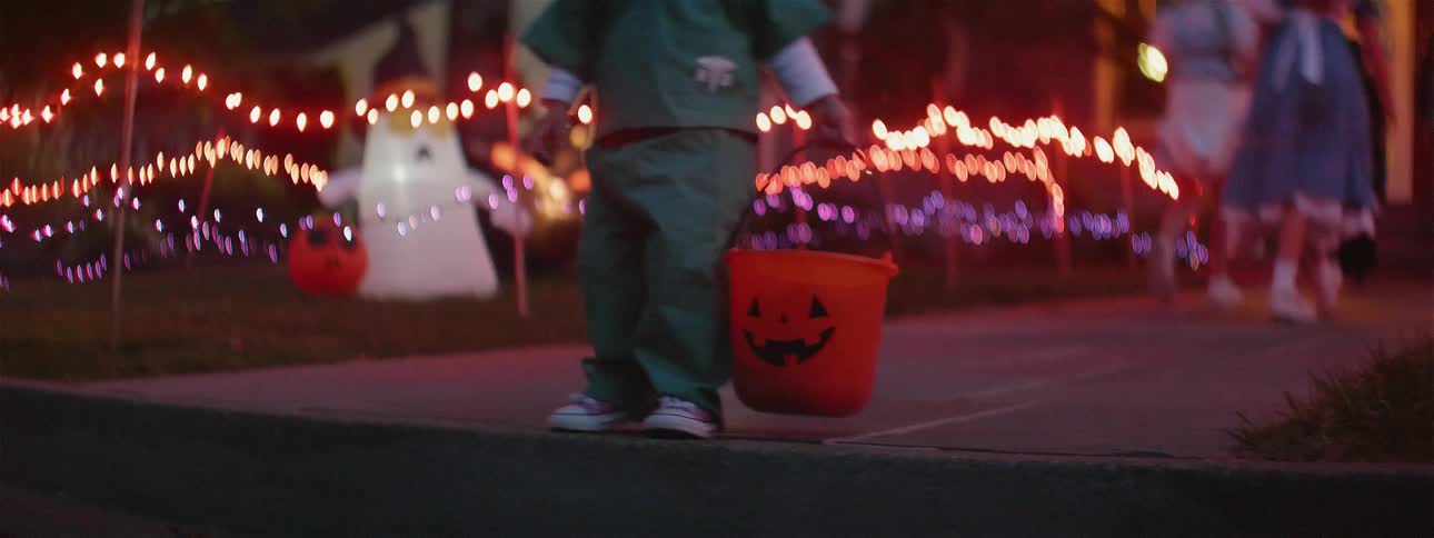 A toddler is carrying a bucket as he trick-or-treats on halloween at dusk