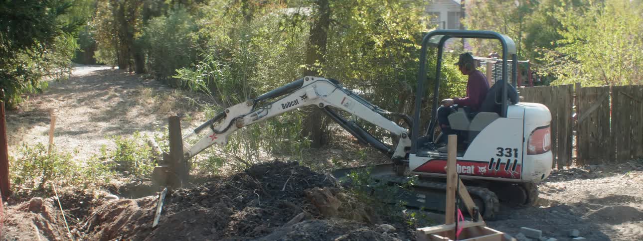 A man uses an excavator to dig around a tree stump