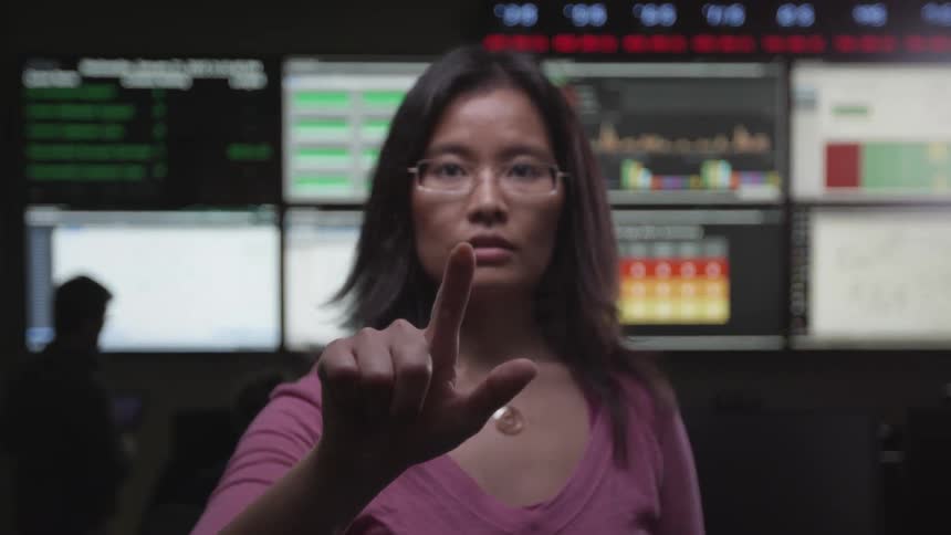 Woman talks in front of monitors displaying charts then points forward and walks away