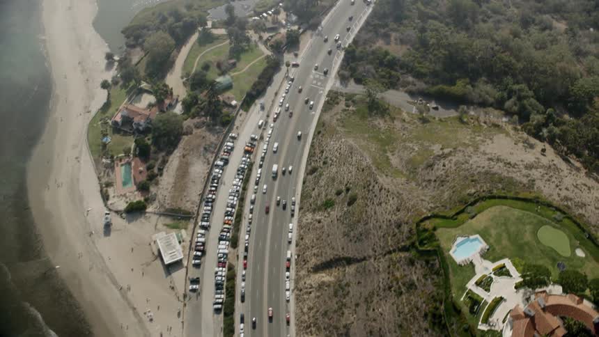 A drone is flying over a coastal highway with waves crashing on the beach