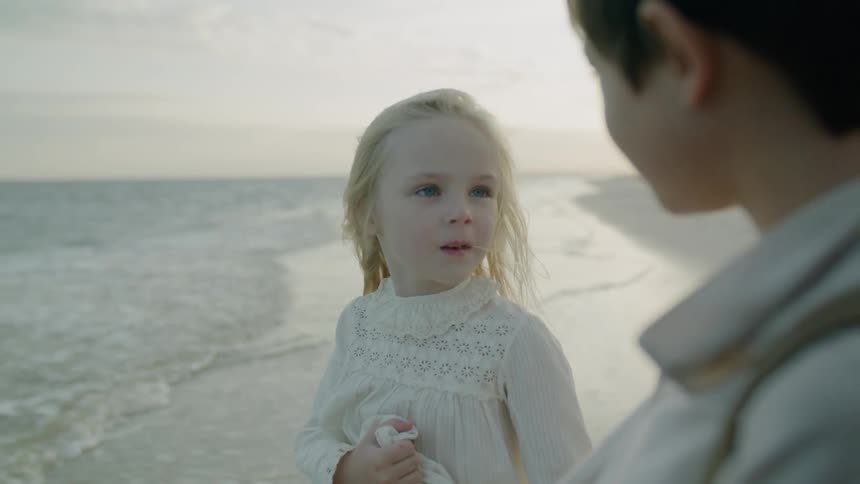A little girl is looking at her brother as they stand at the beach on a windy day