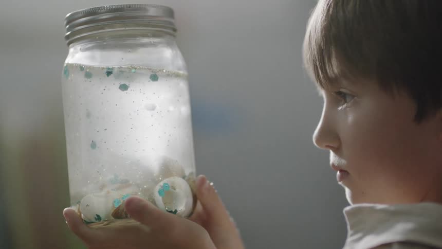 A boy is holding a jar filled with water, glitter, and shells inside during the day