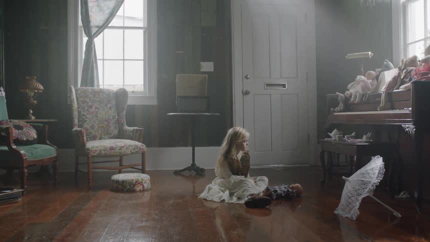 Young blond girl sitting on the floor in a room and looking up at a collection of dolls
