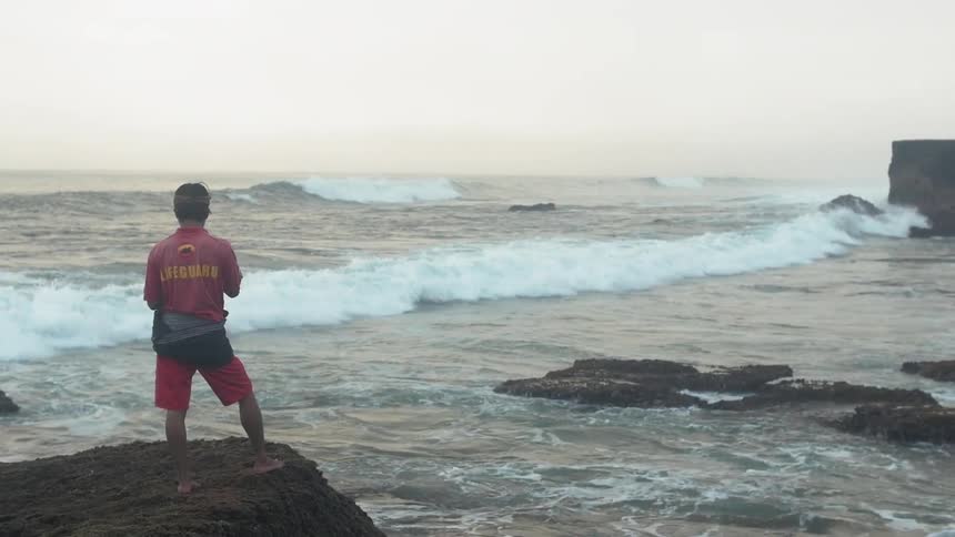 A man is standing on the edge of some rocks next to the ocean as waves crash onto the beach