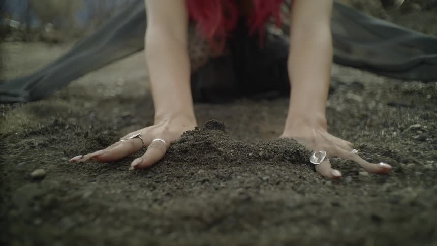 A woman kneels in the dirt and runs her hands over it while her dress blows in the breeze