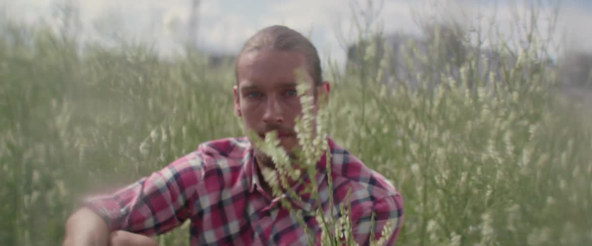 A man sits in overgrown weeds with a large building in the background and clouds in the sky