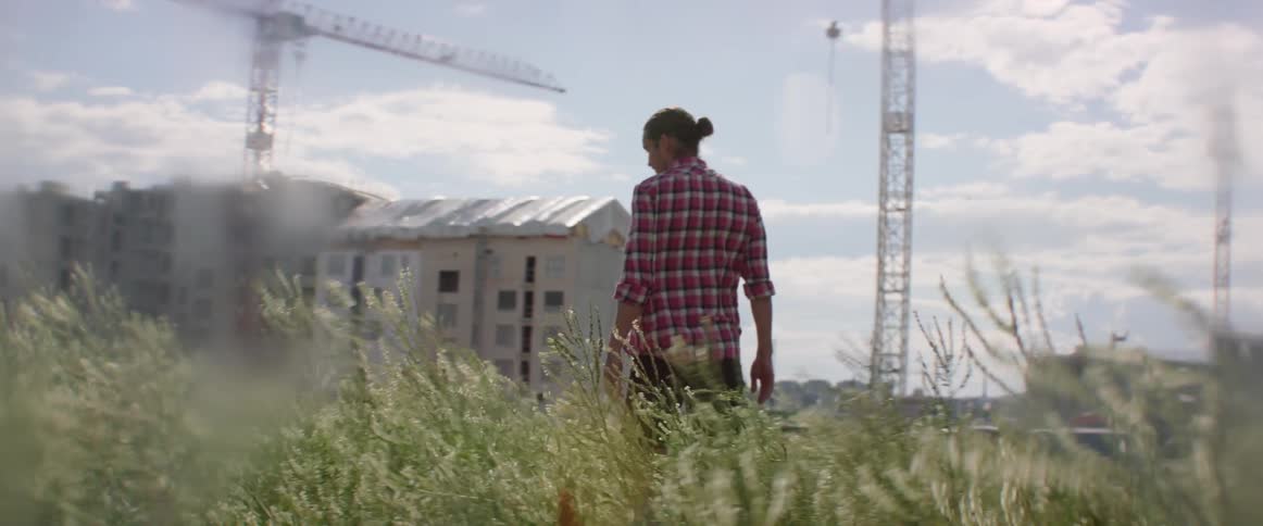 A man is walking in an open field on the outskirts of a construction site on a windy day