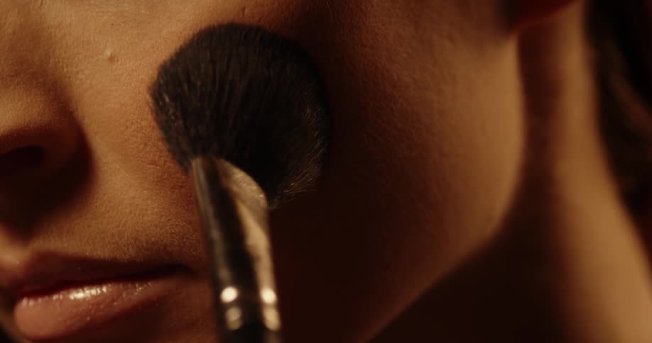 A woman slowly applies blush to her face in the dark