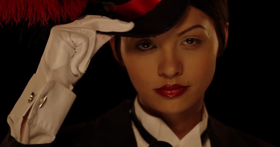 A woman dressed in a suit and a small hat with a feather slowly looks up at the camera while tipping her hat