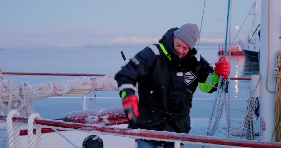 A man is using a mallet to break ice off a ship's railings at dawn