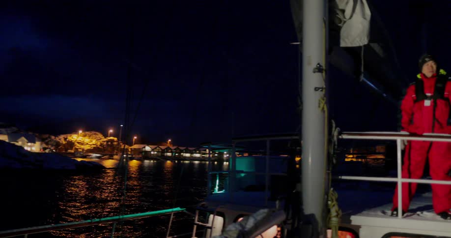 A man is standing at a boat railing as it pulls away from a harbor at night