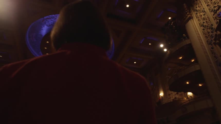 A woman stands on the stage of an empty theater and looks up and around