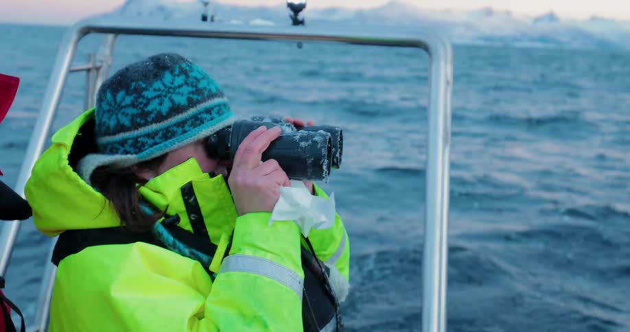 A woman is cleaning her binoculars as she stands in a dinghy on a winter day