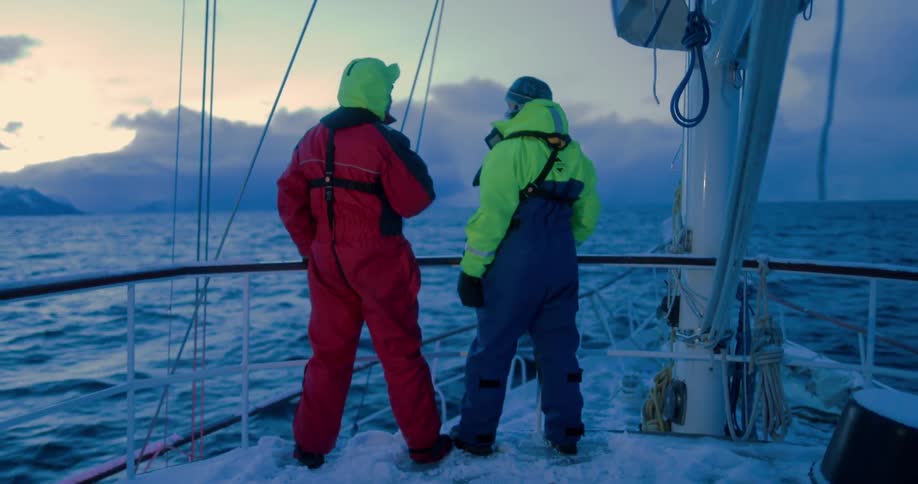 Two men stand on a boat talking as it sails through the ocean at sunset on a cloudy evening