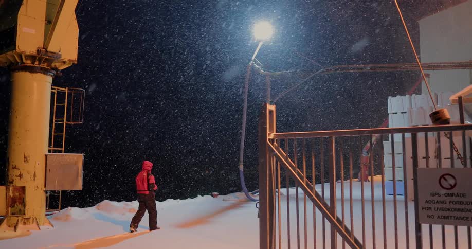 A man is walking on the grounds of an industrial plant outside at night in the snow