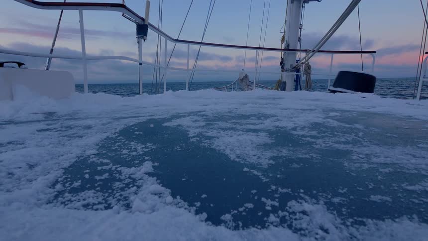 Ice and snow are clinging to the deck of a sailboat at it floats in the ocean at dusk