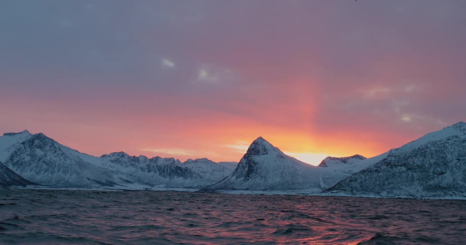 Snow covered mountains at the edge of the ocean with the sun setting behind them