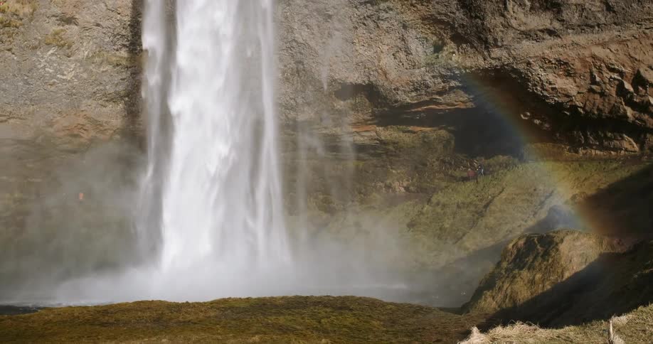 A rainbow is shining at the base of a waterfall on a sunny day
